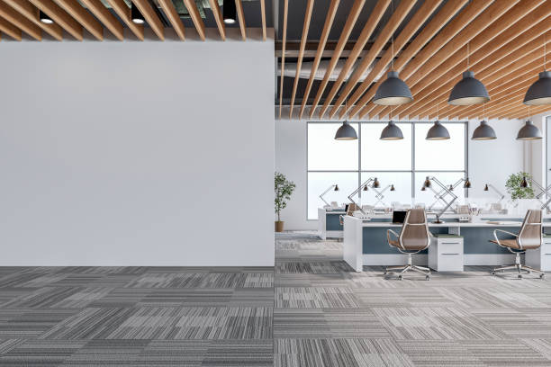 A modern office interior with an open floor plan. On the left, there is a blank white wall with copy space, and on the right, rows of workstations (white and gray) with computers, chairs, and greenery are illuminated by large windows and pendant lights. The ceiling features wooden slats, complementing the gray patterned carpet flooring. 3D rendered image.